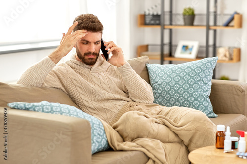 health, cold and people concept - sick young man in blanket with smartphone and medicine calling doctor at home © Syda Productions