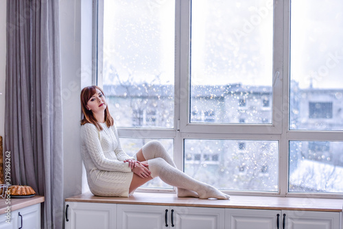 A beautiful young woman in a white knitted sweater dress and long leggings, sits on a wide windowsill at the window. Looking into the distance, sad, loneliness concept. Copy space. White interiors Free Download A beautiful young woman in a white knitted sweater dress and long leggings, sits on a wide windowsill at the window. Looking into the distance, sad, loneliness concept. Copy space. White interiors - GFXLoad