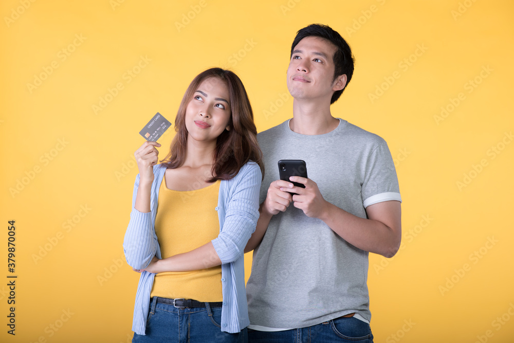 Young Asian couple holding credit card and smartphone while looking up isolated on yellow background Young Asian couple holding credit card and smartphone while looking up isolated on yellow background