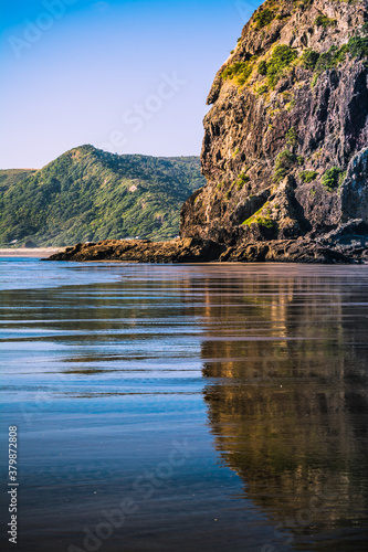Lion Rock reflected in the mirror of black sand at Piha Beach, New Zealand