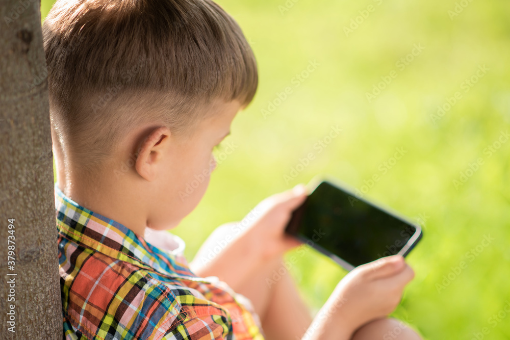 A beautiful child sits on the grass and looks at the phone in the summer at sunset. Cute boy with mobile in hand A beautiful child sits on the grass and looks at the phone in the summer at sunset. Cute boy with mobile in hand
