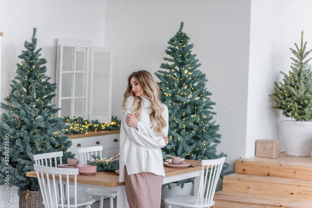 Beautiful curly blonde with long hair by the Christmas trees with garlands of lights. A cheerful girl smiles, dressed in a gray knitted sweater with a large collar, powdery dress, sits in the kitchen Beautiful curly blonde with long hair by the Christmas trees with garlands of lights. A cheerful girl smiles, dressed in a gray knitted sweater with a large collar, powdery dress, sits in the kitchen