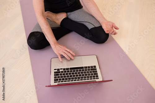 A woman is sitting in a yoga pose on a mat with a laptop. Wooden floor. Daylight. Healthy lifestyle, online classes at home, online shopping, sportswear concept.