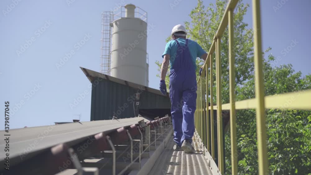 Back view of male Caucasian employee walking along loading conveyor outdoors on sunny day. Wide shot of professional worker controlling equipment on plant or factory. Back view of male Caucasian employee walking along loading conveyor outdoors on sunny day. Wide shot of professional worker controlling equipment on plant or factory.