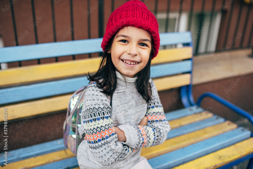 A happy little girl in a red winter hat is sitting on a colorful bench outside and waiting for her friend. Pretty kid takes a rest outdoor after school.