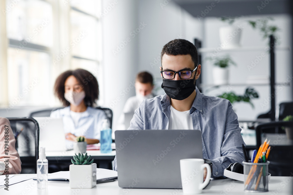 Office center workers are protected from virus outbreak during covid-19 epidemic. Young hipster man in glasses and protective mask works at laptop Office center workers are protected from virus outbreak during covid-19 epidemic. Young hipster man in glasses and protective mask works at laptop