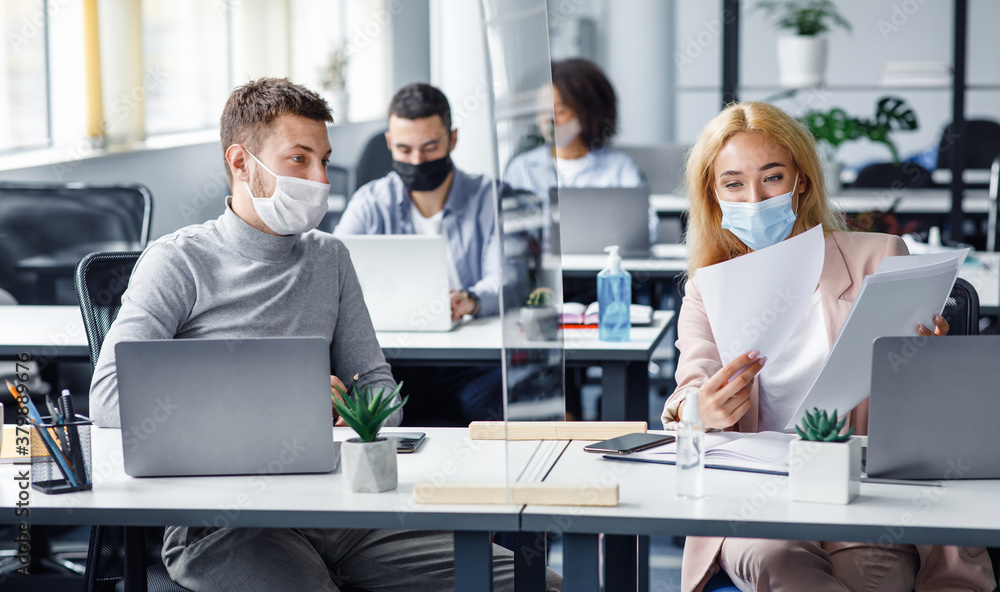 Social distancing and document management. Millennial guy and woman in protective masks discuss work issues through protective board Social distancing and document management. Millennial guy and woman in protective masks discuss work issues through protective board