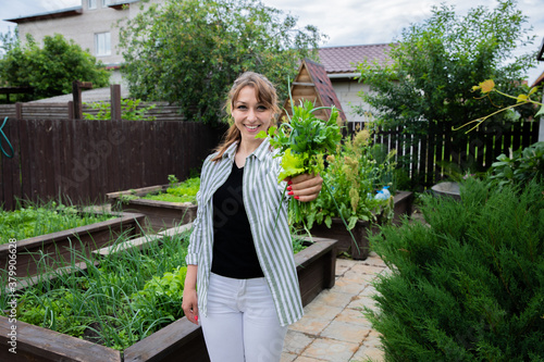 Young beautiful farmer holding fresh harvest herbs greens on garden background, happy woman gardener look at camera with smile, green leek lettuce parsley dill in hands, home organic products, farming