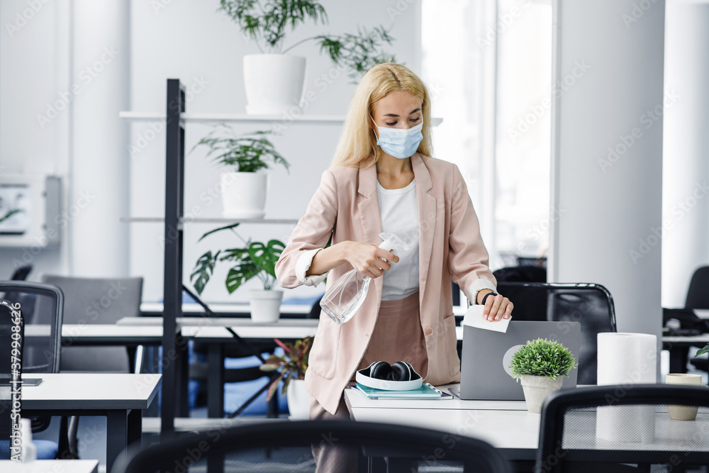 Hygiene and protection of workplace from virus. Attractive millennial woman in protective mask disinfects laptop in office interior Hygiene and protection of workplace from virus. Attractive millennial woman in protective mask disinfects laptop in office interior