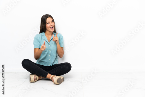 Teenager girl sitting on the floor pointing to the front and smiling