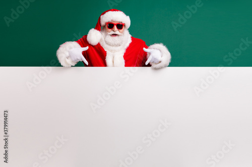 Photo of retired stylish grandfather grey hair beard direct finger white empty board offer present wear red santa x-mas costume coat gloves sunglass headwear isolated green color background