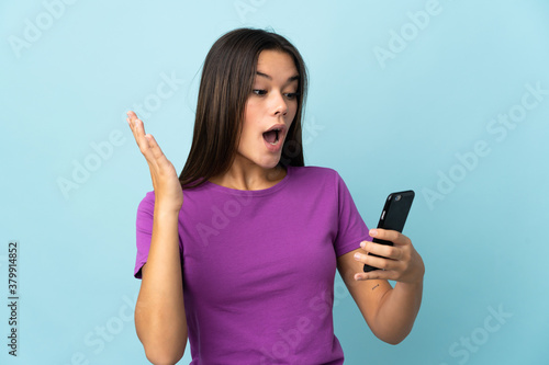 Teenager girl isolated on pink background looking at the camera while using the mobile with surprised expression