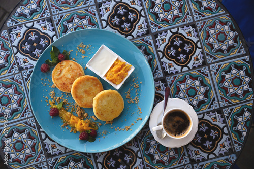 Beautiful ruddy three cheese pancakes on a blue plate on a bright restaurant table with orange peel, mint and raspberries next to cup of coffee. Top view © svitlini
