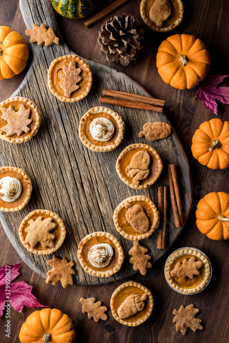 Top down view of various pumpkin pie tarts on a wooden platter, surrounded by autumn decorations.
