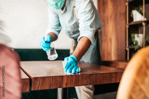 Handsome middle age waiter cleaning and disinfecting restaurant table for next customer. Corona virus and small business is open for work concept.