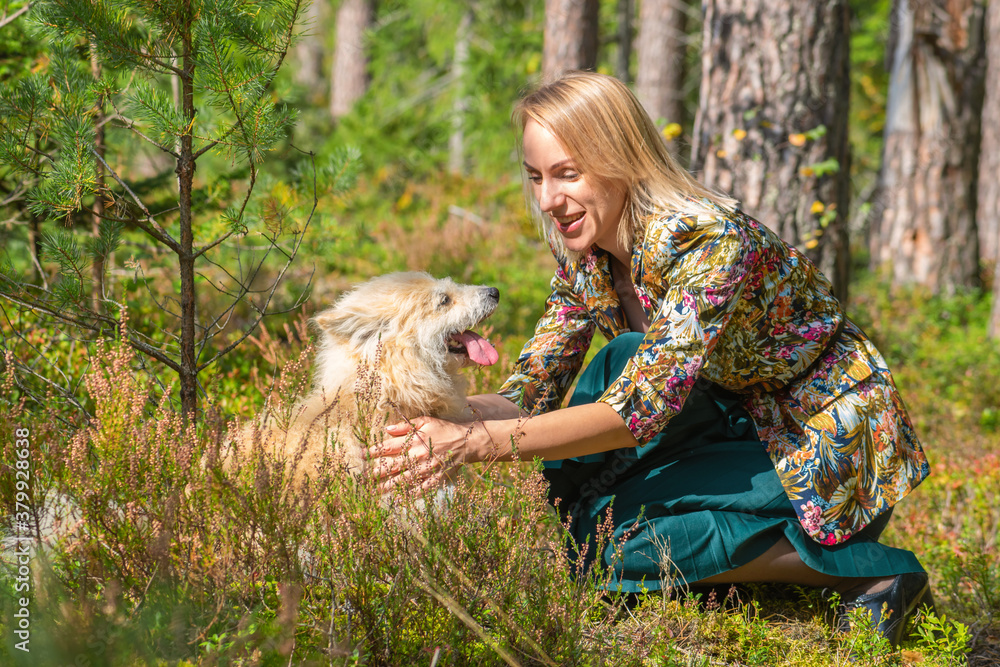 Blonde woman with colorful blouse and green dress playing with beige long haired dog in forest during early autumn time Blonde woman with colorful blouse and green dress playing with beige long haired dog in forest during early autumn time