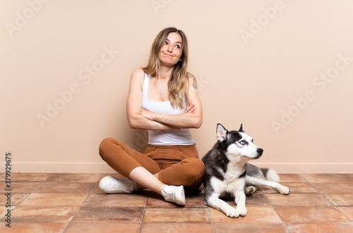 Young pretty woman with her husky dog sitting in the floor at indoors making doubts gesture while lifting the shoulders