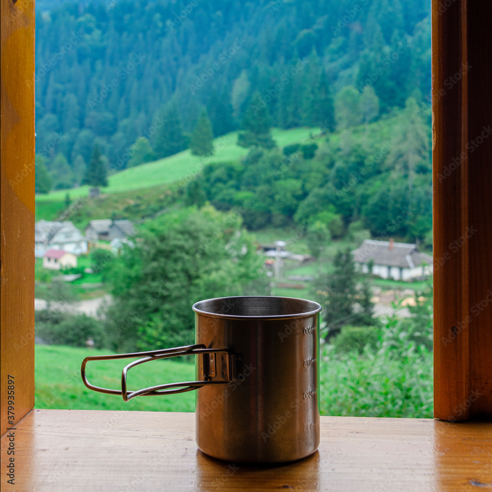 Metal cup of tea stands on a wooden window sill against the backdrop of the mountains