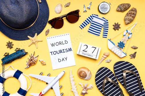 Happy world tourism day. Touristic clothes, hat, flip-flops, sunglasses and decorative items on light background. Flat lay, top view. Calendar date 27 September, notebook with text WORLD TOURISM DAY