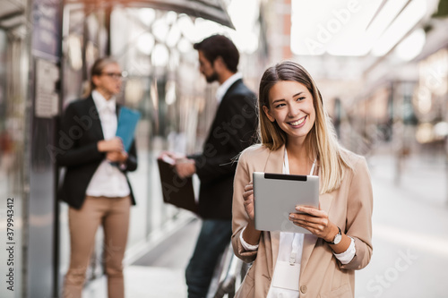 Young happy businesswoman holding digital tablet outside of modern building, businesspeople in background.