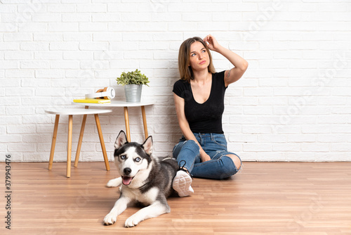 Young pretty woman with her husky dog sitting in the floor at indoors having doubts and with confuse face expression
