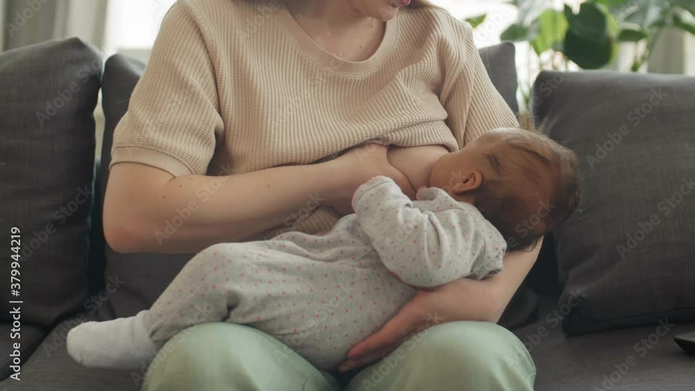 Lockdown of unrecognizable Caucasian woman sitting on couch in living room and breastfeeding her infant daughter lying on her knees Lockdown of unrecognizable Caucasian woman sitting on couch in living room and breastfeeding her infant daughter lying on her knees