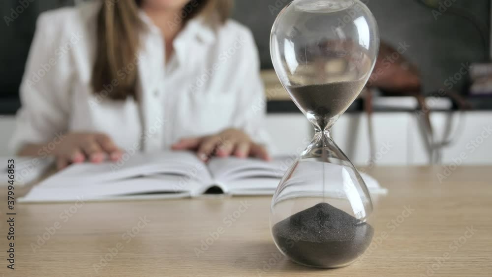 Pouring sand in an hourglass standing on the desk in workspace. Woman reading books at blur background Distance education concept 4k Pouring sand in an hourglass standing on the desk in workspace. Woman reading books at blur background Distance education concept 4k