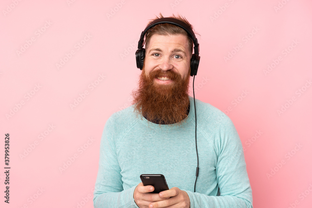 Redhead man with long beard over isolated pink background listening music with a mobile and looking front Redhead man with long beard over isolated pink background listening music with a mobile and looking front