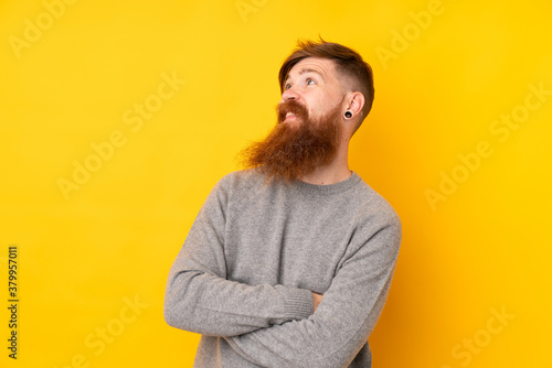 Redhead man with long beard over isolated yellow background looking up while smiling