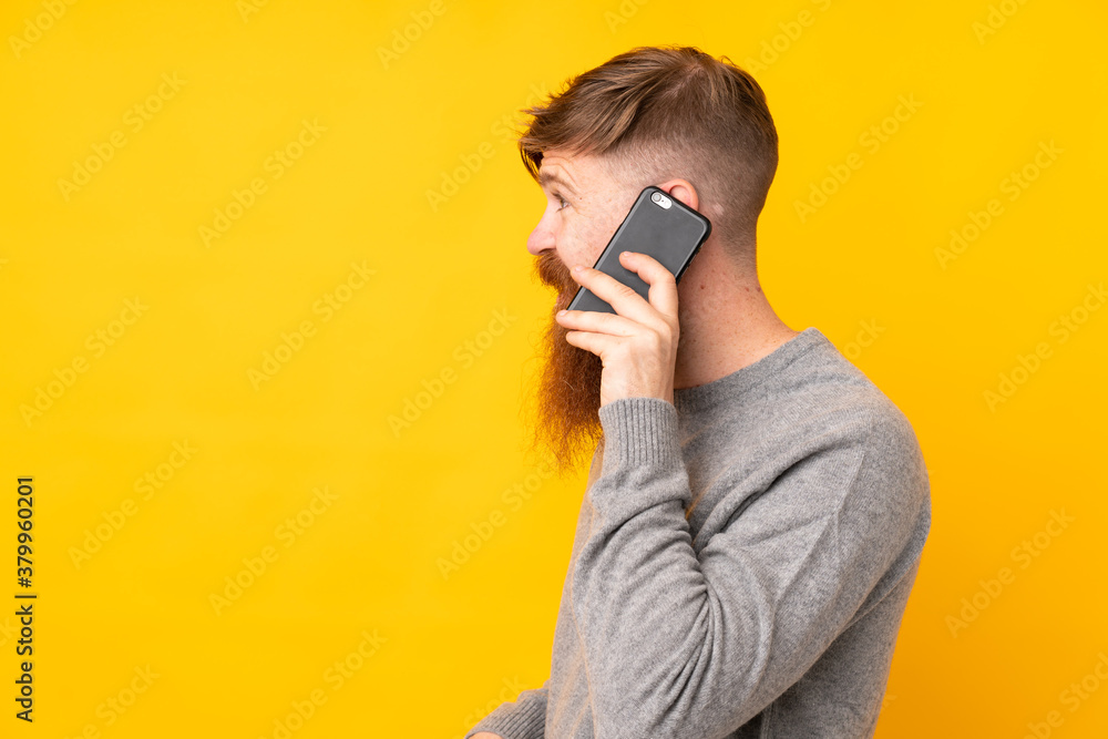 Redhead man with long beard over isolated yellow background keeping a conversation with the mobile phone with someone Redhead man with long beard over isolated yellow background keeping a conversation with the mobile phone with someone
