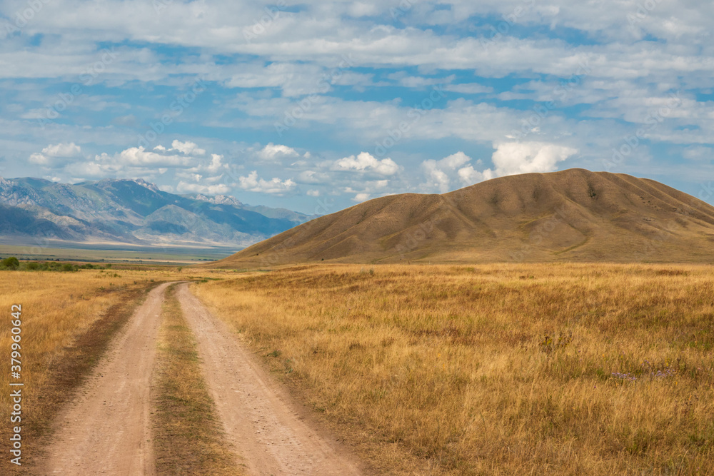 Road heading through Tekes river valley in Almaty Region. Cloudy sky, mountains on the background Road heading through Tekes river valley in Almaty Region. Cloudy sky, mountains on the background