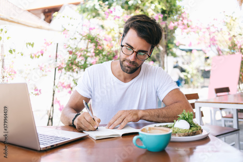 Concentrated young male freelancer organizing project making notes for business planning working remotely on cafe terrace  caucasian creative man blogger sitting at lunch break writing in notebook