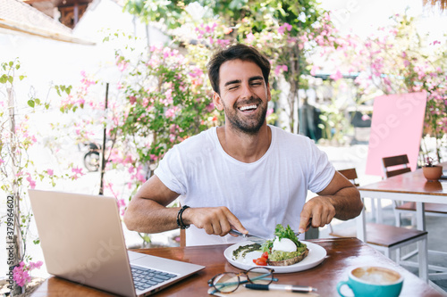 Half length portrait of cheerful caucasian male eating healthy meal satisfied with brunch laughing in good mood  positive funny hipster guy keeping healthy lifestyle enjoying vegan food in cafe