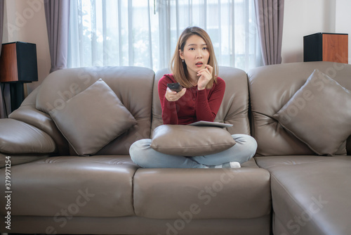 A beautiful woman is sitting on the sofa and pressing the remote control to change the channel as needed. But she was surprised with what she was seeing.