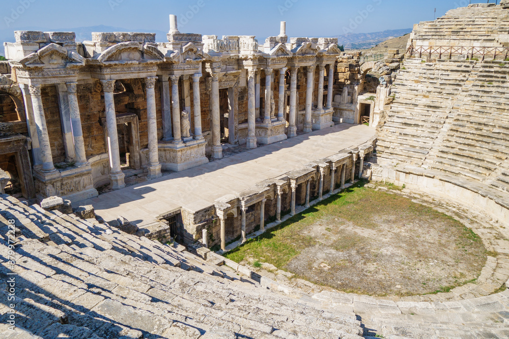 Side view onto antique theater in ancient city Hierapolis, Pamukkale, Turkey. There are empty audience with lines of seats, & scene with classic decorations: statues, reliefs, columns, portico etc