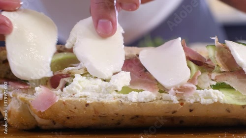 Young chef puts thin slices of mozzarella cheese on sandwich in kitchen on cutting Board in home interior. Preparation of morning Breakfast from bun cucumber cheese ham. Baguette sandwich.