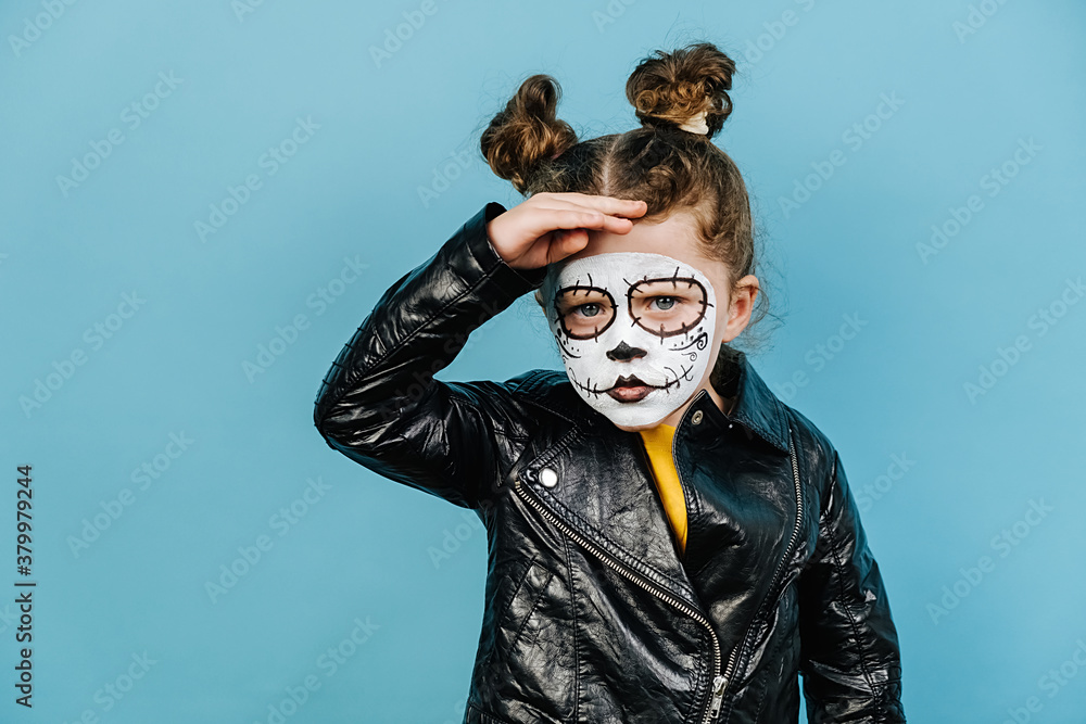 Angry little girl with serious expression, focused into distance, keeps hand on forehead, wears creative makeup, isolated over blue background, dressed in Halloween costume. Mexico city's Day of Dead