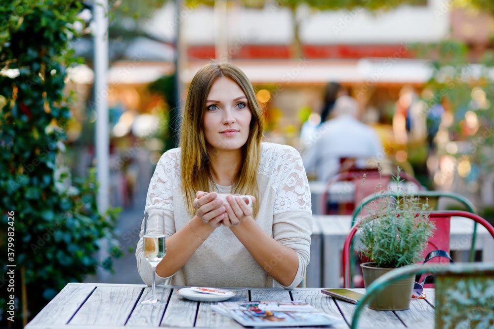 Fototapeta premium Young beautiful woman drinking cappuccino and cup of coffee and glass champagne while waiting for friend and breakfast. Happy alone woman in outdoor cafe or restaurant on sunny summer day.