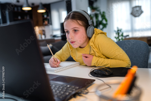 Cute child with headphones using laptop computer for online learning. Home school. Girl doing homework at home. Lifestyle concept for home schooling. Social distancing and education. photo