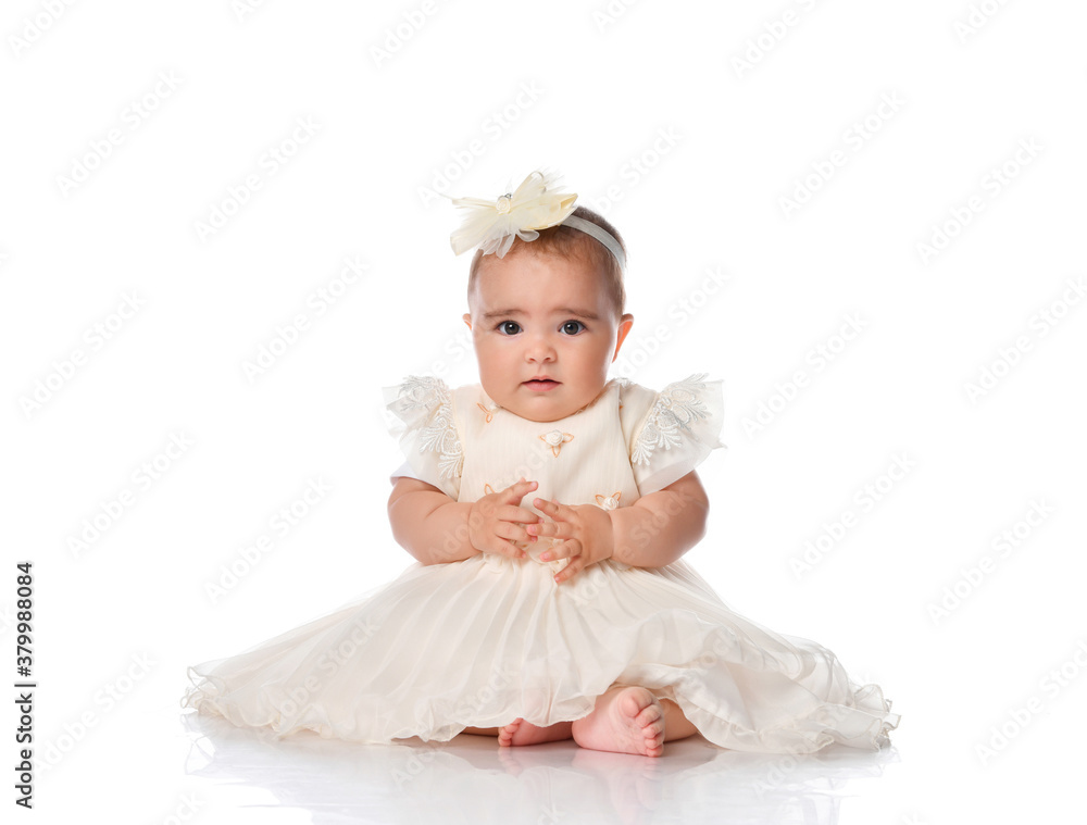 Beautiful baby in a festive dress and a hair band sitting on the floor. Toddler toddler looking at camera studio portrait shot on white background. Children's fashion and beauty Beautiful baby in a festive dress and a hair band sitting on the floor. Toddler toddler looking at camera studio portrait shot on white background. Children's fashion and beauty
