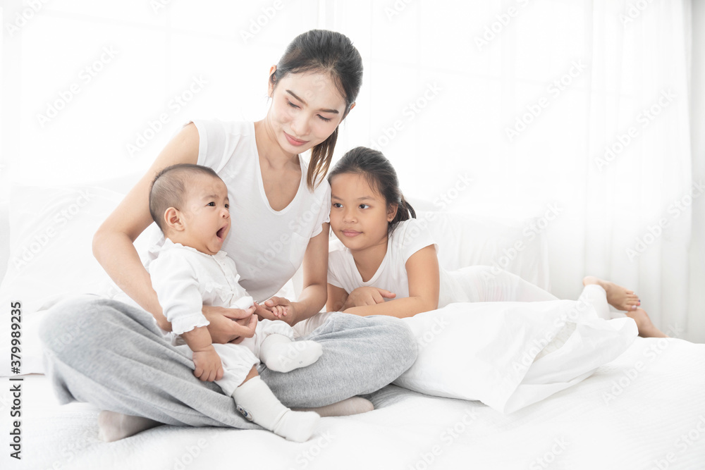 Asian mother and children girl baby laugh together at home. Asia mom holdind infant and play with sister sitting on white bed in living room at house family having fun together. Asian mother and children girl baby laugh together at home. Asia mom holdind infant and play with sister sitting on white bed in living room at house family having fun together.