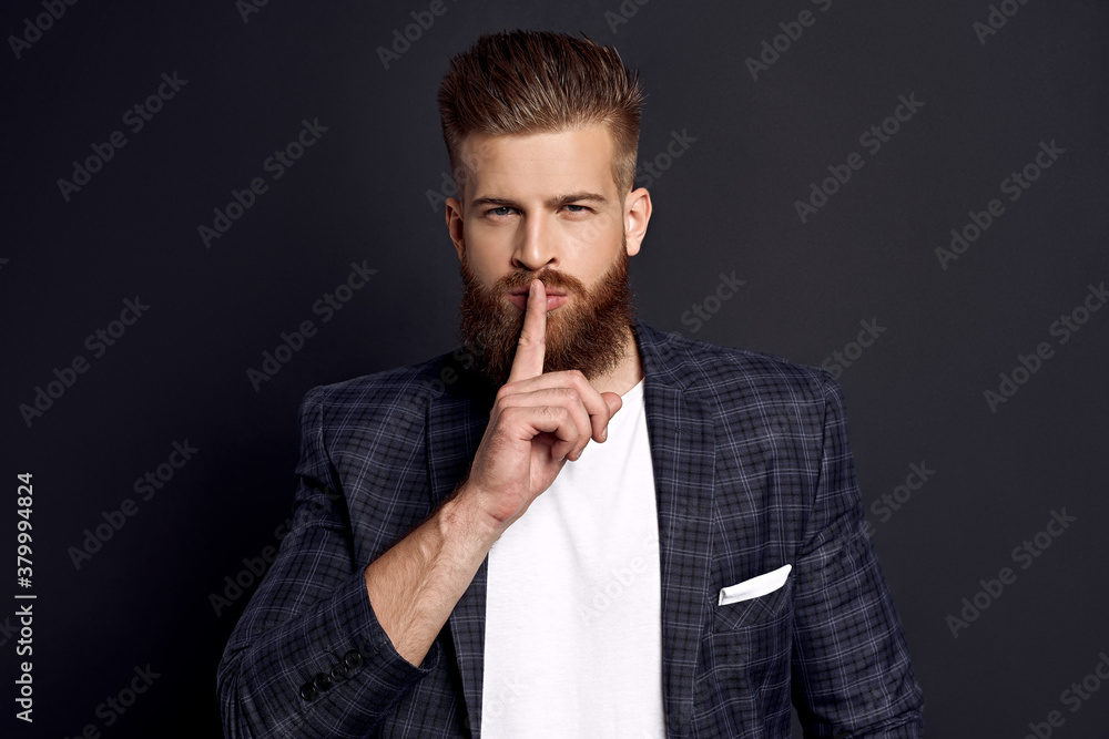 Handsome bearded man in suit is asking for silence. He is raising finger to his lips. The man is standing and looking at the camera secretly. Isolated on black background Handsome bearded man in suit is asking for silence. He is raising finger to his lips. The man is standing and looking at the camera secretly. Isolated on black background