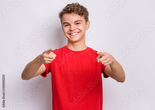 Portrait of happy teen boy pointing fingers at camera. Cute smiling child in red t-shirt choose you, on grey background. Laughing child having fan.
