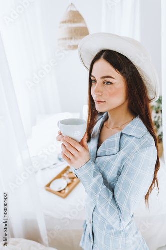 Beautiful young stylish woman in business suit and white hat standing near bed and drink coffee in sunny morning.