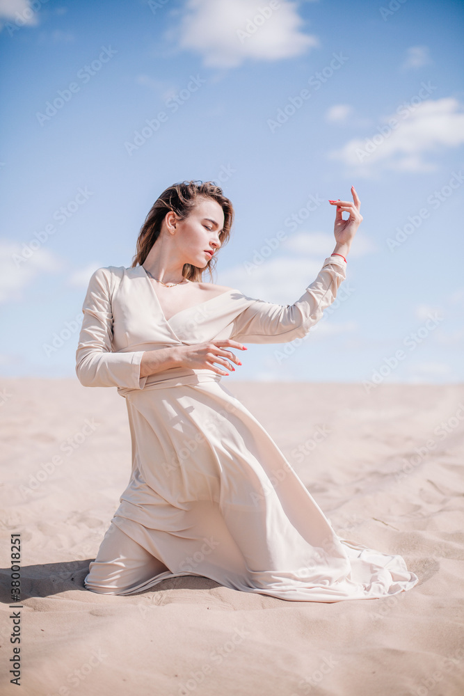 A young, slender girl in a beige dress poses in the wind in the desert