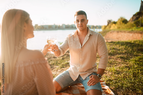 A cute blonde woman in a pastel pink dress and a caucasian man drinking white wine on the beach at sunset. A loving couple celebrating and enjoying a picnic on the river bank
