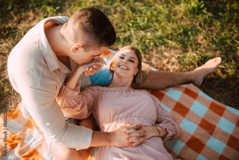 Fototapeta premium A beautiful blonde caucasian woman in a dress is lying on a blanket next to her man. Romantic loving couple on a picnic by the river