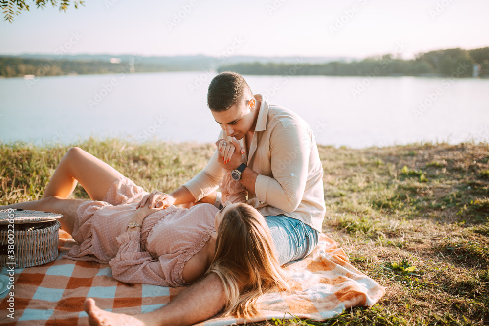 Fototapeta premium A beautiful blonde caucasian woman in a dress is lying on a blanket next to her man. Romantic loving couple on a picnic by the river