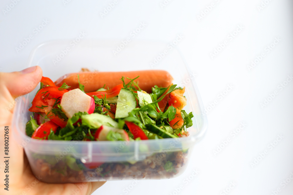 Tasty appetizing food in open plastic transparent container, held by hand of young Caucasian man on white background. Fresh vegetable salad, sausage and buckwheat. Cooking for outdoor event. Tasty appetizing food in open plastic transparent container, held by hand of young Caucasian man on white background. Fresh vegetable salad, sausage and buckwheat. Cooking for outdoor event.