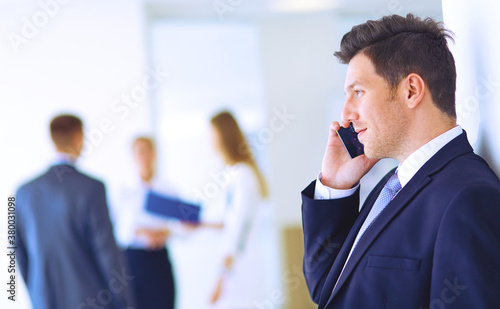 Portrait of young businessman in office with colleagues in the background and using mobile
